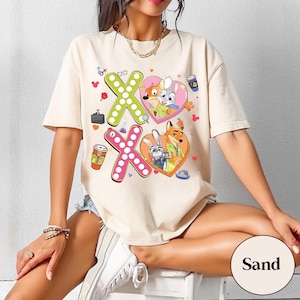 May include: Sand-colored t-shirt featuring a graphic design with the characters from Zootopia. The design includes two heart-shaped images with the characters Nick Wilde and Judy Hopps, along with the letters "XO" in green and pink.