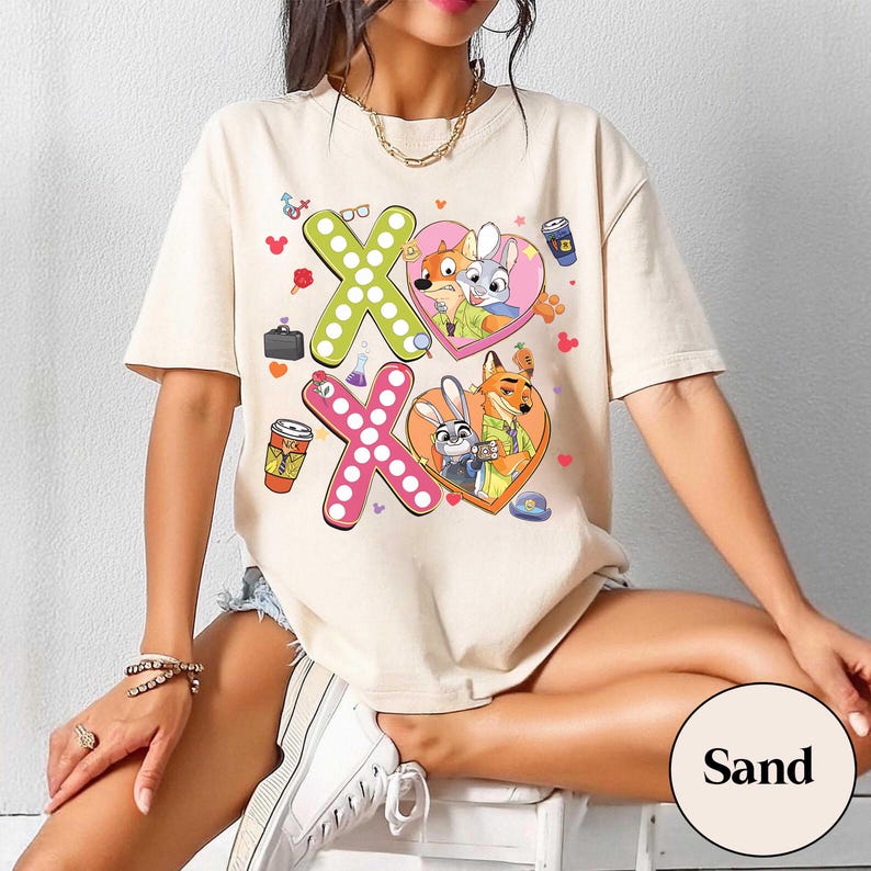 May include: Sand-colored t-shirt featuring a graphic design with the characters from Zootopia. The design includes two heart-shaped images with the characters Nick Wilde and Judy Hopps, along with the letters "XO" in green and pink.