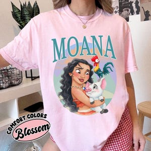 May include: A light pink t-shirt with the word "MOANA" in teal letters. The shirt features a graphic of Moana holding a pig, with a rooster on top. The words "COMFORT COLORS" and "Blossom" are printed on the bottom left.