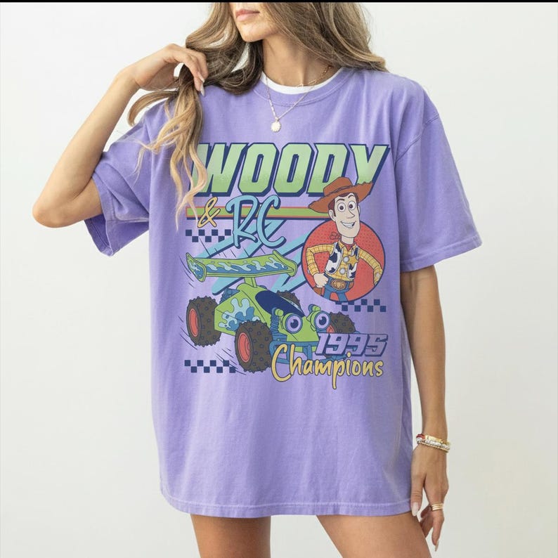 May include: A lilac-colored t-shirt featuring a graphic of Woody and RC from Toy Story. The design includes the text "WOODY & RC" in green and blue, a cartoon image of Woody, and a green and blue RC car. The text "1995 Champions" is also visible.