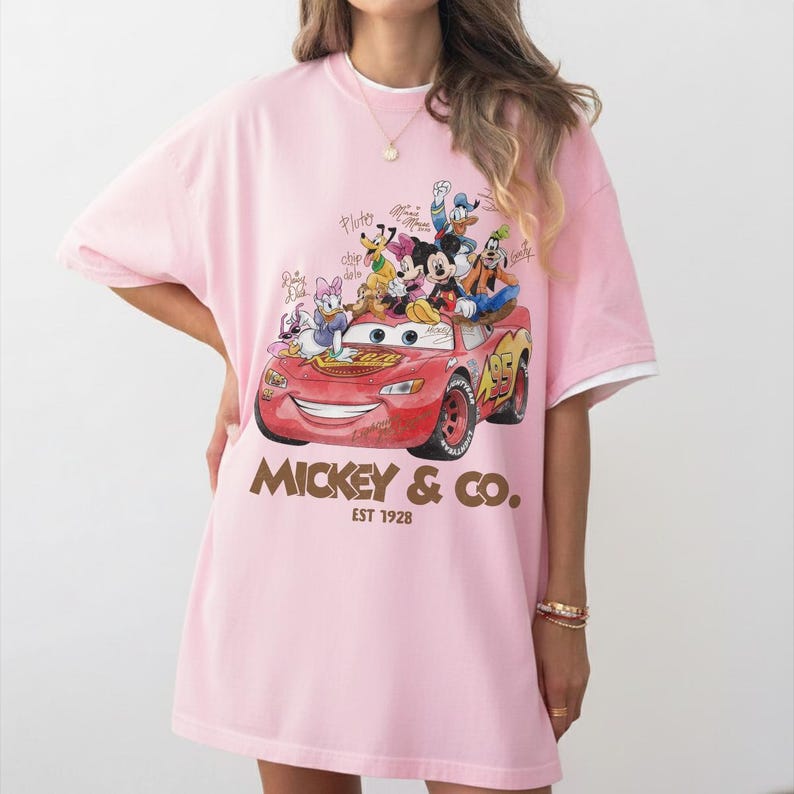 May include: Pink t-shirt featuring a red car with Disney characters, including Mickey Mouse, Donald Duck, and Daisy Duck. The shirt has the text "Mickey & Co. EST 1928" in brown. The shirt has white trim on the sleeves.