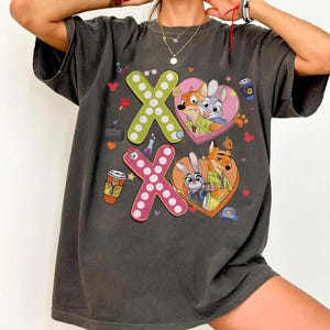 May include: Dark gray t-shirt featuring the characters from Zootopia. The shirt has a design with the letters X and O, with the characters inside heart shapes. The design includes coffee cups and other small graphics.