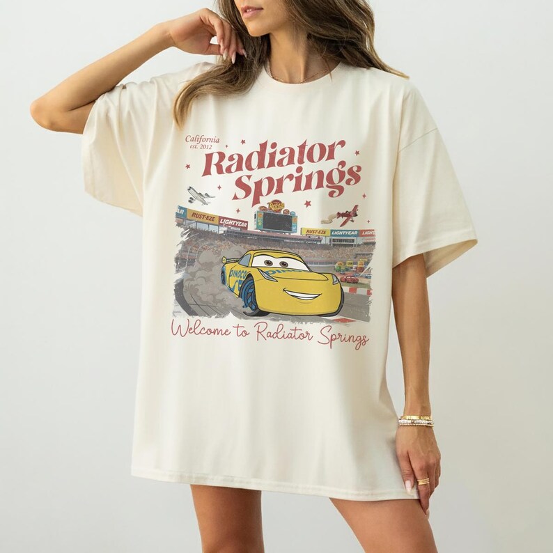 May include: Cream-colored t-shirt featuring a graphic of Radiator Springs from the movie Cars. The design includes the text "Radiator Springs" and "Welcome to Radiator Springs", along with an image of the character Lightning McQueen.