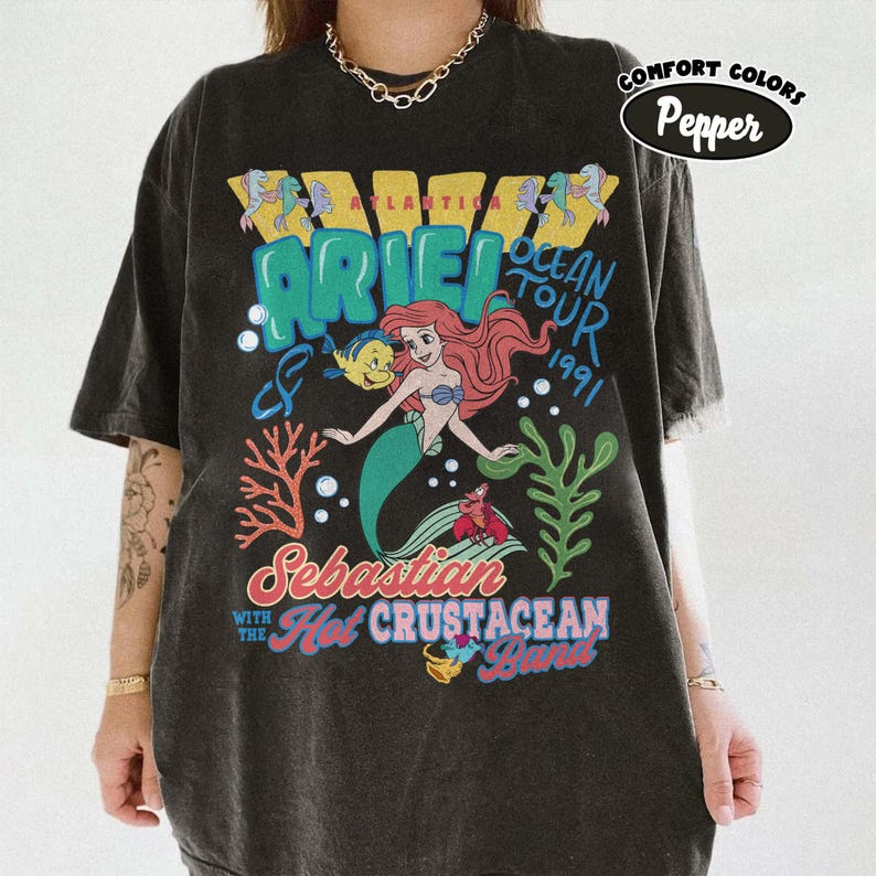 May include: A charcoal gray t-shirt featuring a graphic print of Ariel, Flounder, and Sebastian. The text reads Atlantica Ariel Ocean Tour 1991, Sebastian with the Hot Crustacean Band. A Comfort Colors Pepper label is visible in the upper right corner.