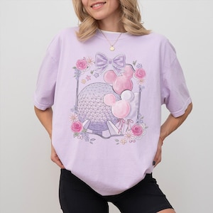 K&ouml;nnte beinhalten: Ein helllila T-Shirt mit einem grafischen Design mit einer Schleife, Mickey Mouse-Ballons und einer geometrischen Kugel, umrahmt von Blumen. Das Shirt besteht aus einem weichen, bequemen Material.