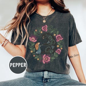 May include: Dark gray t-shirt featuring a floral wreath design with a bear and a figure. The wreath includes pink and purple flowers, green leaves, and yellow accents. The word "PEPPER" is printed on a circular label.