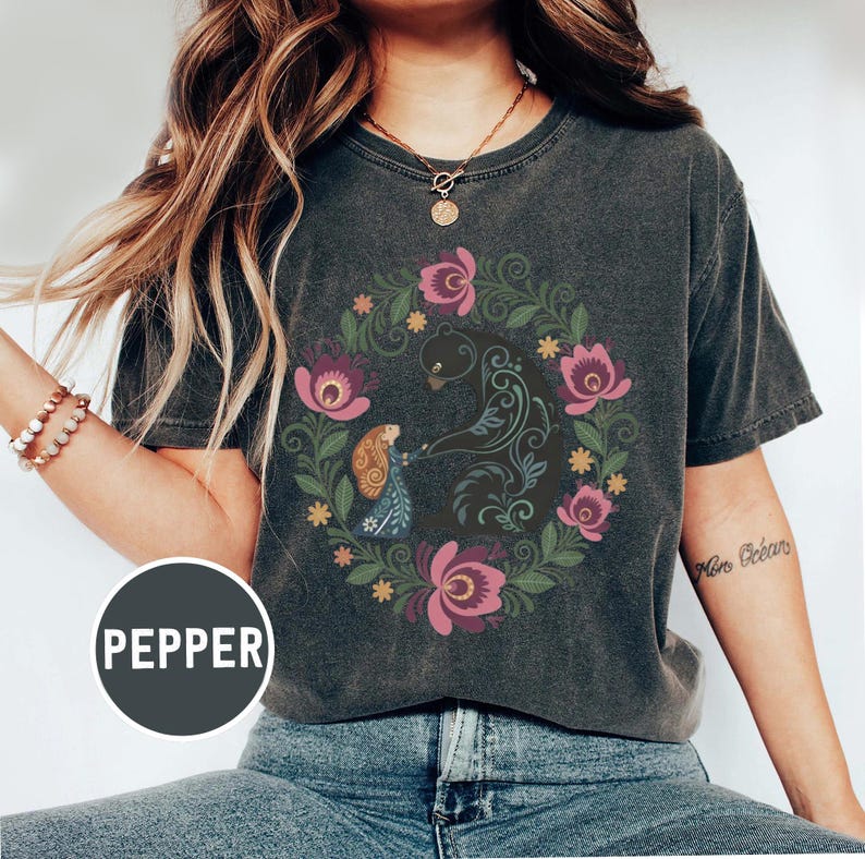 May include: Dark gray t-shirt featuring a floral wreath design with a bear and a figure. The wreath includes pink and purple flowers, green leaves, and yellow accents. The word "PEPPER" is printed on a circular label.
