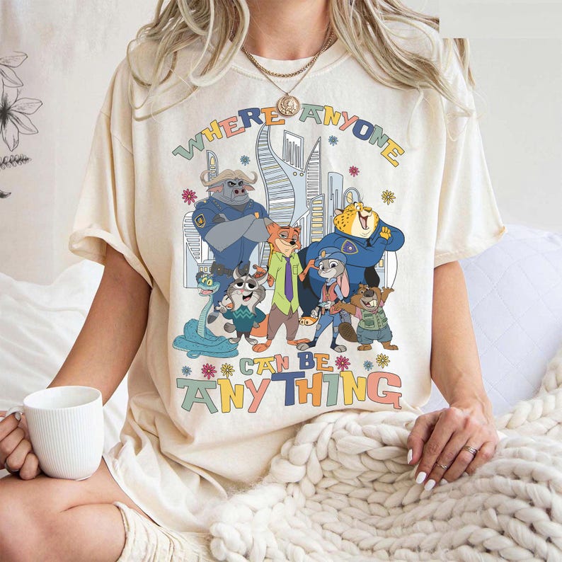 Puede incluir: Camiseta color crema con un gr&aacute;fico colorido de personajes de dibujos animados y el texto "WHERE ANYONE CAN BE ANYTHING". El dise&ntilde;o incluye un paisaje urbano y acentos florales.