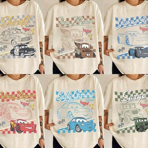 May include: Cream-coloured t-shirts with cartoon car designs, including "Sheriff," "Tow Mater," and "Jackson Storm." Each shirt displays a different car graphic, text, and the year "2006" or "2005."