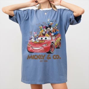 May include: Blue t-shirt featuring a graphic of Lightning McQueen from the movie Cars, with Mickey Mouse and friends. The graphic includes the text "Mickey & Co. EST 1928". The shirt has a relaxed fit and short sleeves.