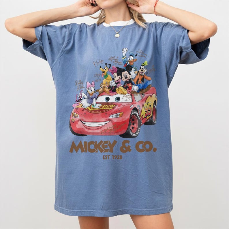 May include: Blue t-shirt featuring a graphic of Lightning McQueen from the movie Cars, with Mickey Mouse and friends. The graphic includes the text "Mickey & Co. EST 1928". The shirt has a relaxed fit and short sleeves.