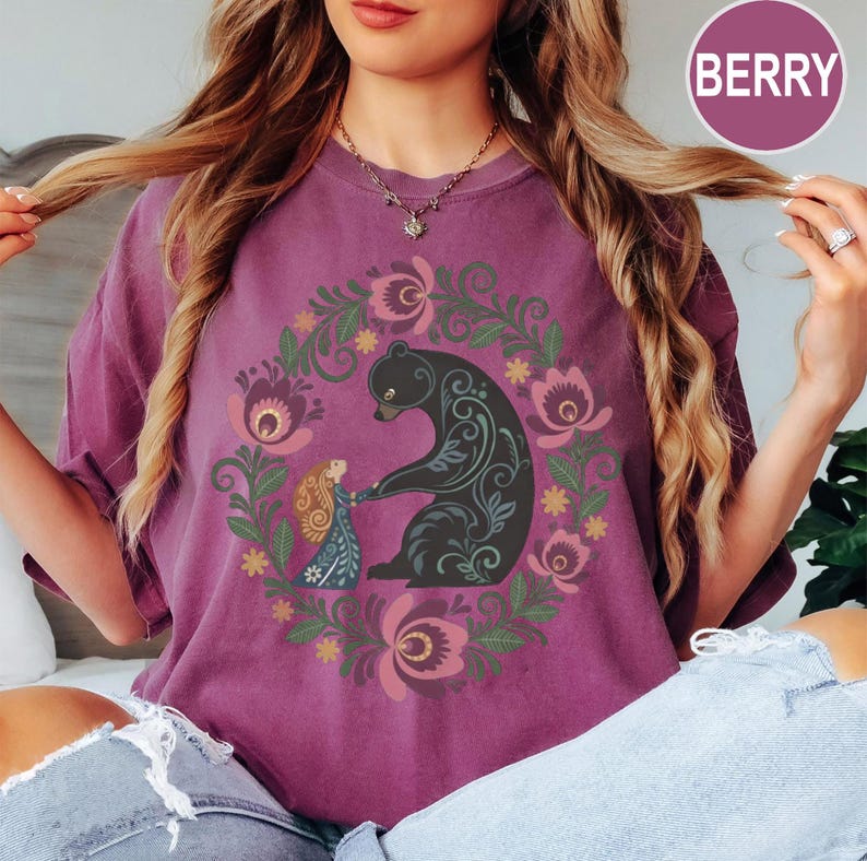 May include: Berry-colored t-shirt featuring a circular floral design with a black bear and a girl holding hands. The bear has decorative swirls, and the girl wears a blue dress. The design is surrounded by pink and green flowers.