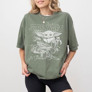 Puede incluir: Una camiseta verde oliva con un estampado gr&aacute;fico en blanco de Grogu, una nave espacial y el texto Star Wars, This is the Way y The Mandalorian. El dise&ntilde;o incluye un icono de casco estilizado y un elemento de patr&oacute;n de cuadros al frente.