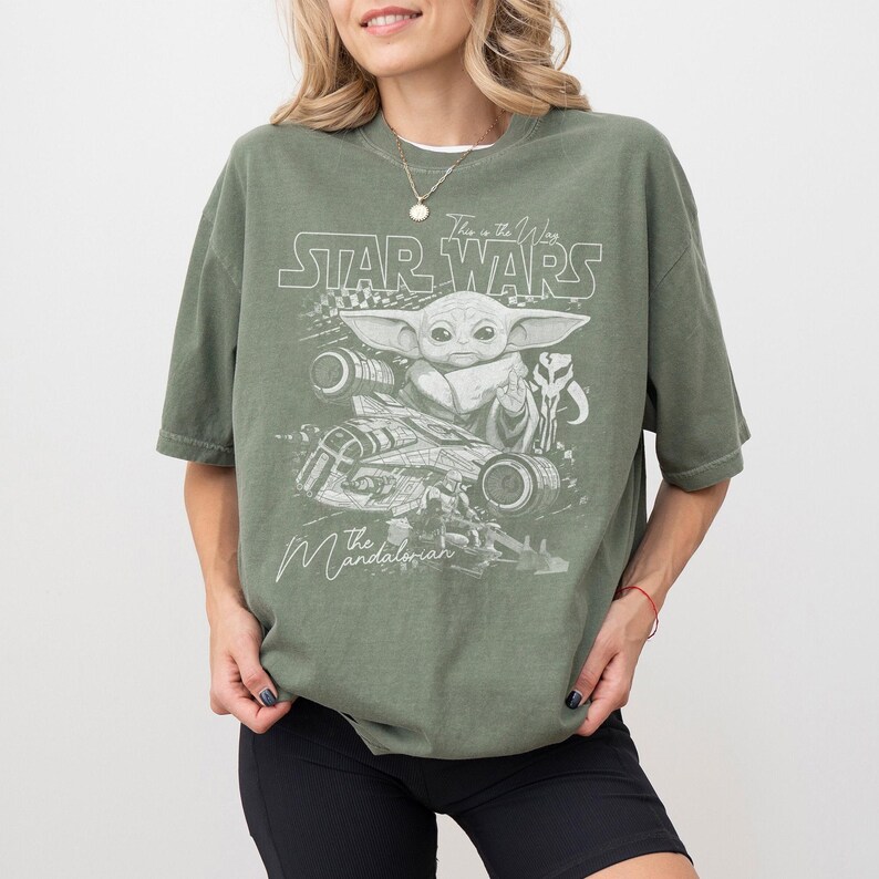 Puede incluir: Una camiseta verde oliva con un estampado gr&aacute;fico en blanco de Grogu, una nave espacial y el texto Star Wars, This is the Way y The Mandalorian. El dise&ntilde;o incluye un icono de casco estilizado y un elemento de patr&oacute;n de cuadros al frente.