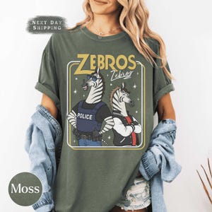 May include: Moss green t-shirt with a cartoon graphic of two zebras. One zebra is in a police vest, the other in a letterman jacket. The text "ZEBROS" and "Zebros" is displayed above the zebras.