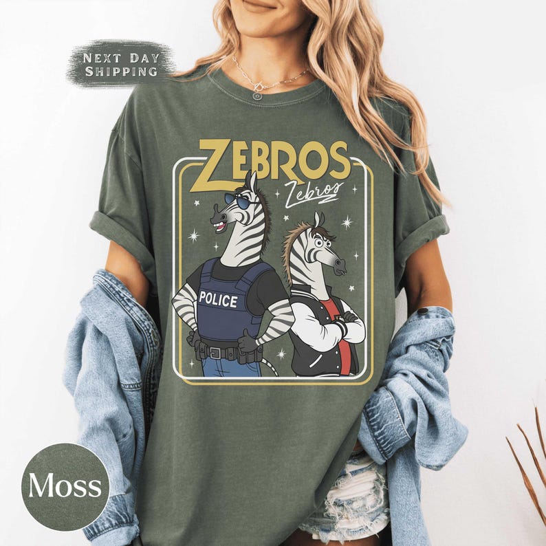 May include: Moss green t-shirt with a cartoon graphic of two zebras. One zebra is in a police vest, the other in a letterman jacket. The text "ZEBROS" and "Zebros" is displayed above the zebras.