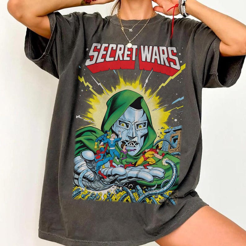 May include: Dark gray t-shirt featuring a comic book illustration with the text "SECRET WARS" in red and white. The graphic depicts a metallic-faced character in a green hood, surrounded by other comic book characters.