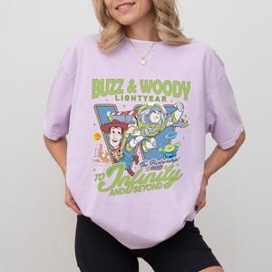 Peut inclure: Un t-shirt &agrave; manches courtes violet clair avec une illustration de Buzz l'&Eacute;clair et Woody de Toy Story. Le texte indique BUZZ & WOODY LIGHTYEAR et TO INFINITY AND BEYOND, avec The Partnership! en &eacute;criture cursive. Le motif inclut des &eacute;toiles.