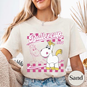 May include: Cream-colored t-shirt featuring a cartoon unicorn named Buttercup with a yellow mane and horn. The design includes the text "Buttercup 1995" in pink, set against a pink and white checkered background.