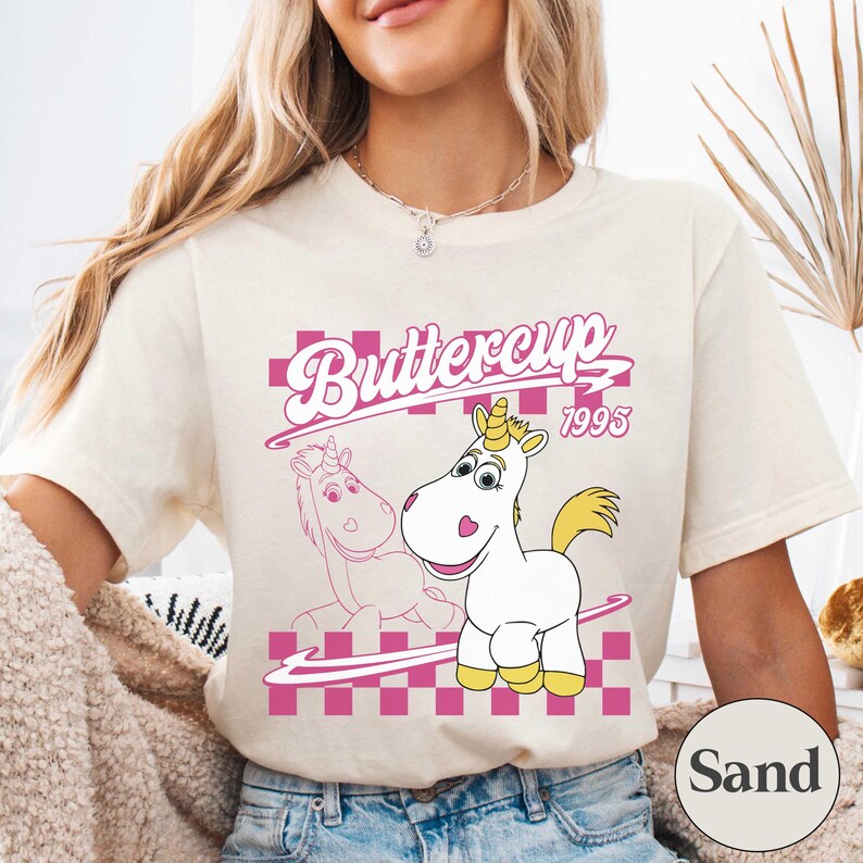 May include: Cream-colored t-shirt featuring a cartoon unicorn named Buttercup with a yellow mane and horn. The design includes the text "Buttercup 1995" in pink, set against a pink and white checkered background.