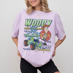 May include: A light purple t-shirt featuring a graphic of Woody from Toy Story with a race car. The text on the shirt reads "Woody & RC 1995 Champions". The design includes green, blue, red, and yellow accents.