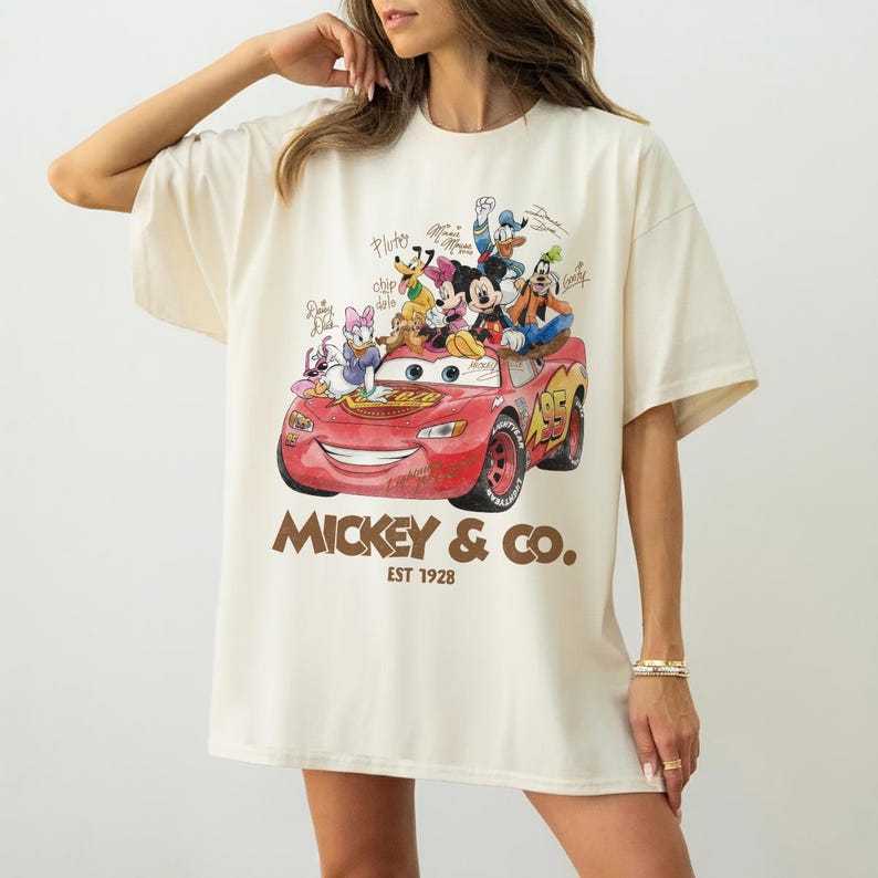 May include: Cream-colored t-shirt featuring a red cartoon car with Disney characters, including Mickey Mouse, Donald Duck, and Goofy. The text "Mickey & Co. EST 1928" is printed below the car. The shirt has short sleeves and a relaxed fit.