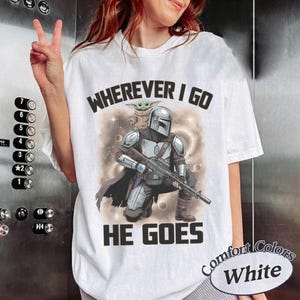 Puede incluir: Una camiseta blanca con un gr&aacute;fico de un personaje blindado con casco que sostiene un rifle con una peque&ntilde;a criatura verde sobre el hombro. El texto de arriba dice WHEREVER I GO y el de abajo HE GOES en letras may&uacute;sculas negras.