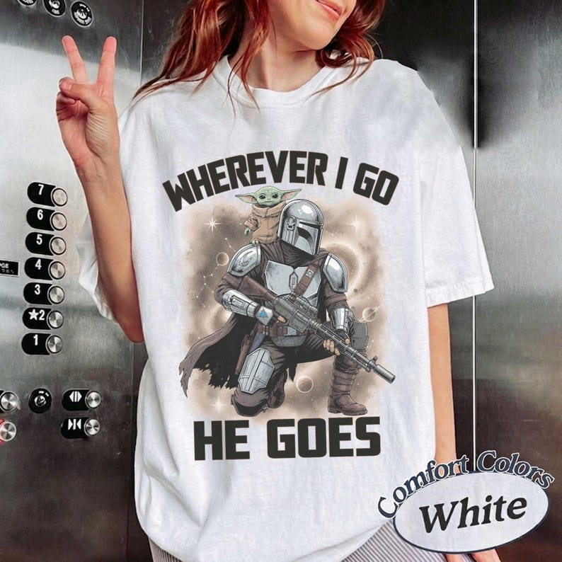 Puede incluir: Una camiseta blanca con un gr&aacute;fico de un personaje blindado con casco que sostiene un rifle con una peque&ntilde;a criatura verde sobre el hombro. El texto de arriba dice WHEREVER I GO y el de abajo HE GOES en letras may&uacute;sculas negras.
