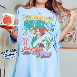 May include: A light blue t-shirt featuring a graphic print of Ariel, Flounder, and Sebastian from The Little Mermaid. The text reads Atlantica Ocean Tour 1991, Sebastian with the Hot Crustacean Band. The design includes coral and seaweed illustrations.