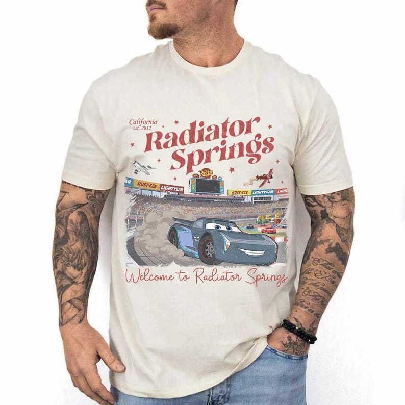 May include: Cream-colored t-shirt featuring a graphic of a race car and the words "Radiator Springs" in red. The shirt also includes the text "California est. 2012" and "Welcome to Radiator Springs".