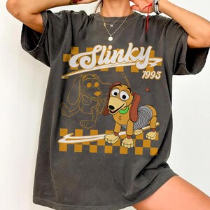 May include: Dark gray t-shirt featuring a cartoon image of Slinky Dog with the text "Slinky 1995". The design includes a yellow and brown checkered background and a line drawing of Slinky Dog.