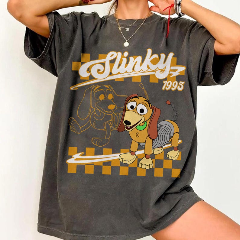 May include: Dark gray t-shirt featuring a cartoon image of Slinky Dog with the text "Slinky 1995". The design includes a yellow and brown checkered background and a line drawing of Slinky Dog.