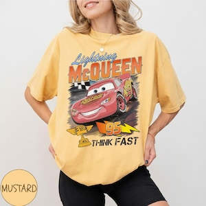 May include: Mustard-colored t-shirt featuring a graphic of Lightning McQueen from the movie Cars. The design includes the text "Lightning McQueen", "95", and "Think Fast". The shirt has short sleeves and a crew neck.