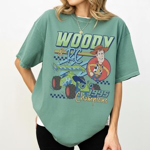 May include: Sage green t-shirt featuring a graphic of Woody and RC from Toy Story. The design includes the text "WOODY & RC" in blue and green, a cartoon image of Woody, and the words "1995 Champions" in yellow.