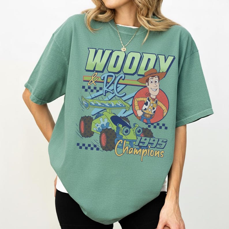 May include: Sage green t-shirt featuring a graphic of Woody and RC from Toy Story. The design includes the text "WOODY & RC" in blue and green, a cartoon image of Woody, and the words "1995 Champions" in yellow.