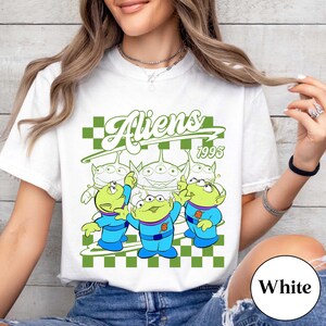 May include: White t-shirt featuring a graphic of three green aliens in blue uniforms, with the word "Aliens" in green script above them. The year "1995" is also displayed. The design is set against a green and white checkered background.