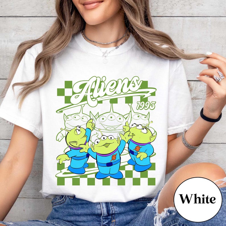May include: White t-shirt featuring a graphic of three green aliens in blue uniforms, with the word "Aliens" in green script above them. The year "1995" is also displayed. The design is set against a green and white checkered background.