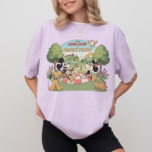 May include: A light purple t-shirt featuring a graphic of Mickey Mouse and Minnie Mouse having a picnic with Pluto. The text "Mickey Mouse Perfect Picnic" is above the characters. The design is in a vintage cartoon style.