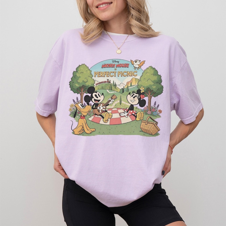 May include: A light purple t-shirt featuring a graphic of Mickey Mouse and Minnie Mouse having a picnic with Pluto. The text "Mickey Mouse Perfect Picnic" is above the characters. The design is in a vintage cartoon style.