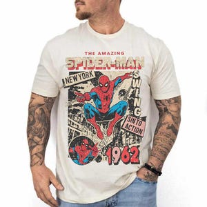 Pode incluir: Camiseta creme com um gr&aacute;fico vintage do Homem-Aranha. O design inclui as palavras "The Amazing Spider-Man", "New York" e "1962". O Homem-Aranha &eacute; retratado em seu cl&aacute;ssico traje vermelho e azul.