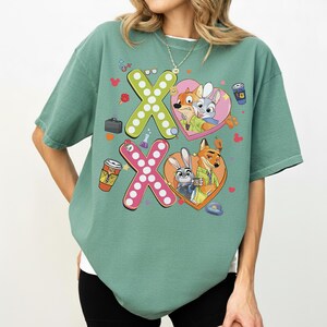 May include: A sage green t-shirt featuring a graphic design with the characters Nick Wilde and Judy Hopps from Zootopia. The design includes the letters "X" and "O" with heart-shaped elements and cartoon images of the characters. The shirt has a relaxed fit.