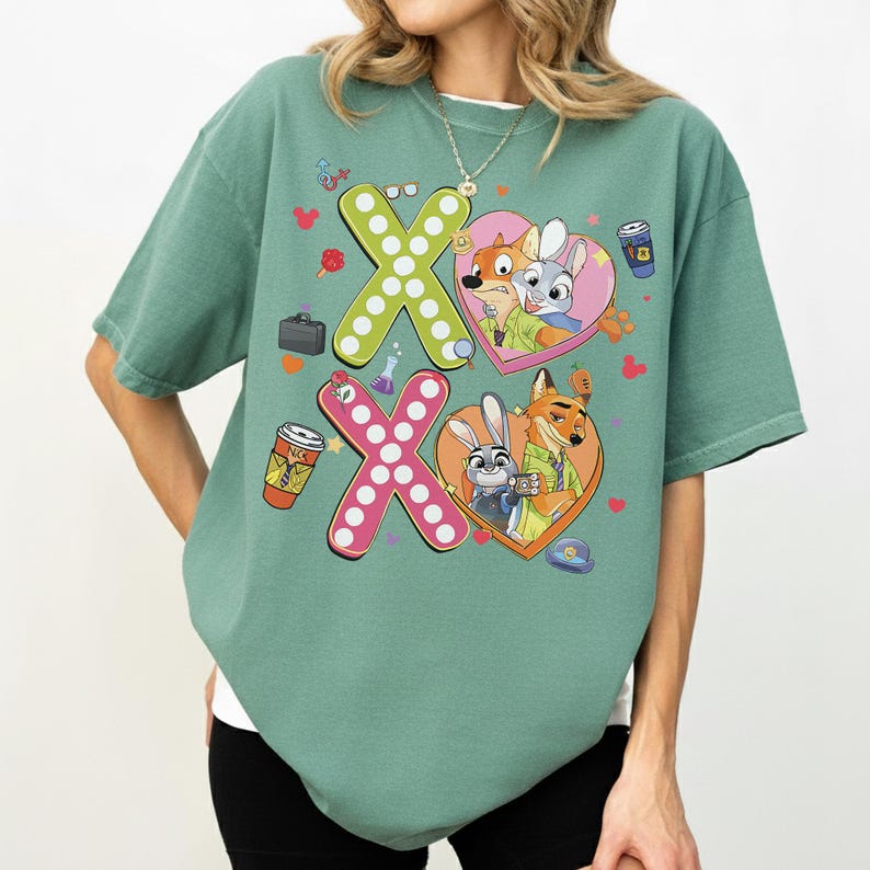 May include: A sage green t-shirt featuring a graphic design with the characters Nick Wilde and Judy Hopps from Zootopia. The design includes the letters "X" and "O" with heart-shaped elements and cartoon images of the characters. The shirt has a relaxed fit.