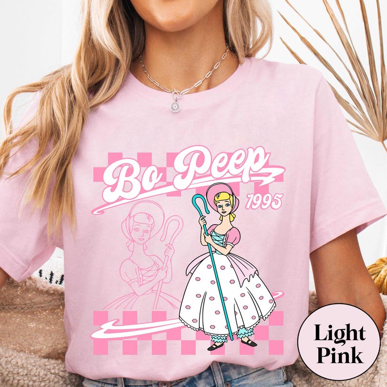 May include: Light pink t-shirt featuring a graphic of Bo Peep from Toy Story, with the text "Bo Peep 1995". The design includes a pink checkered background and a drawing of Bo Peep holding a staff. The shirt is a light pink color.