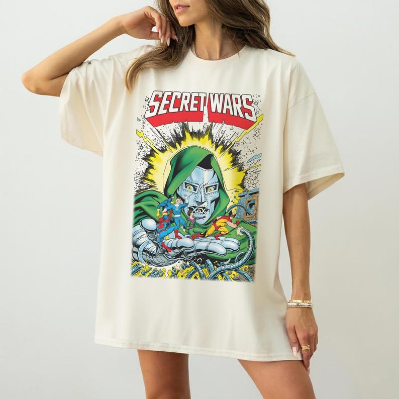 May include: Cream-colored t-shirt featuring a comic book graphic. The graphic includes the text "SECRET WARS" in red and white, along with a depiction of a character in a green hood and silver mask holding other comic book characters.