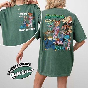 May include: Light green t-shirt featuring cartoon characters from Zootopia. The front of the shirt has the text "Not on our watch" with cartoon characters. The back of the shirt has the text "Zootopia Let's Boogie" with cartoon characters.