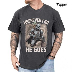 Puede incluir: Una camiseta gris carb&oacute;n de manga corta con un gr&aacute;fico de un guerrero con casco y armadura plateada sosteniendo un rifle, con una peque&ntilde;a criatura verde sobre el hombro. El texto dice WHEREVER I GO HE GOES en letras de color beige claro.