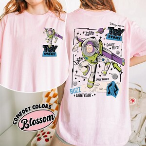 K&ouml;nnte beinhalten: Hellrosa T-Shirt mit einer Grafik von Buzz Lightyear aus Toy Story. Das Design enth&auml;lt den Text "Toy Story", "Buzz + Lightyear" und "Galactic Hero!" im Cartoon-Stil. Das Shirt hat auch den Text "Comfort Colors Blossom".