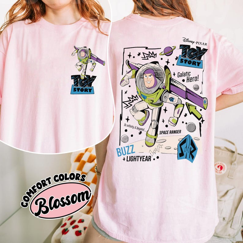K&ouml;nnte beinhalten: Hellrosa T-Shirt mit einer Grafik von Buzz Lightyear aus Toy Story. Das Design enth&auml;lt den Text "Toy Story", "Buzz + Lightyear" und "Galactic Hero!" im Cartoon-Stil. Das Shirt hat auch den Text "Comfort Colors Blossom".