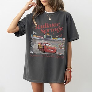May include: Dark gray t-shirt featuring a graphic of Lightning McQueen from the movie Cars on a race track. The text "Radiator Springs" and "Welcome to Radiator Springs" are printed in red.