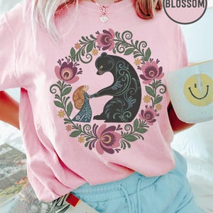 May include: Pink t-shirt featuring a floral wreath design with a black bear and a small figure holding hands. The design includes purple and pink flowers and green leaves. The word "BLOSSOM" is in a pink circle.